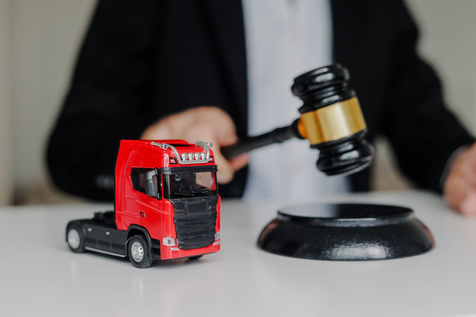 A person holding a judge’s gavel behind a small red toy semi-truck on a desk.
