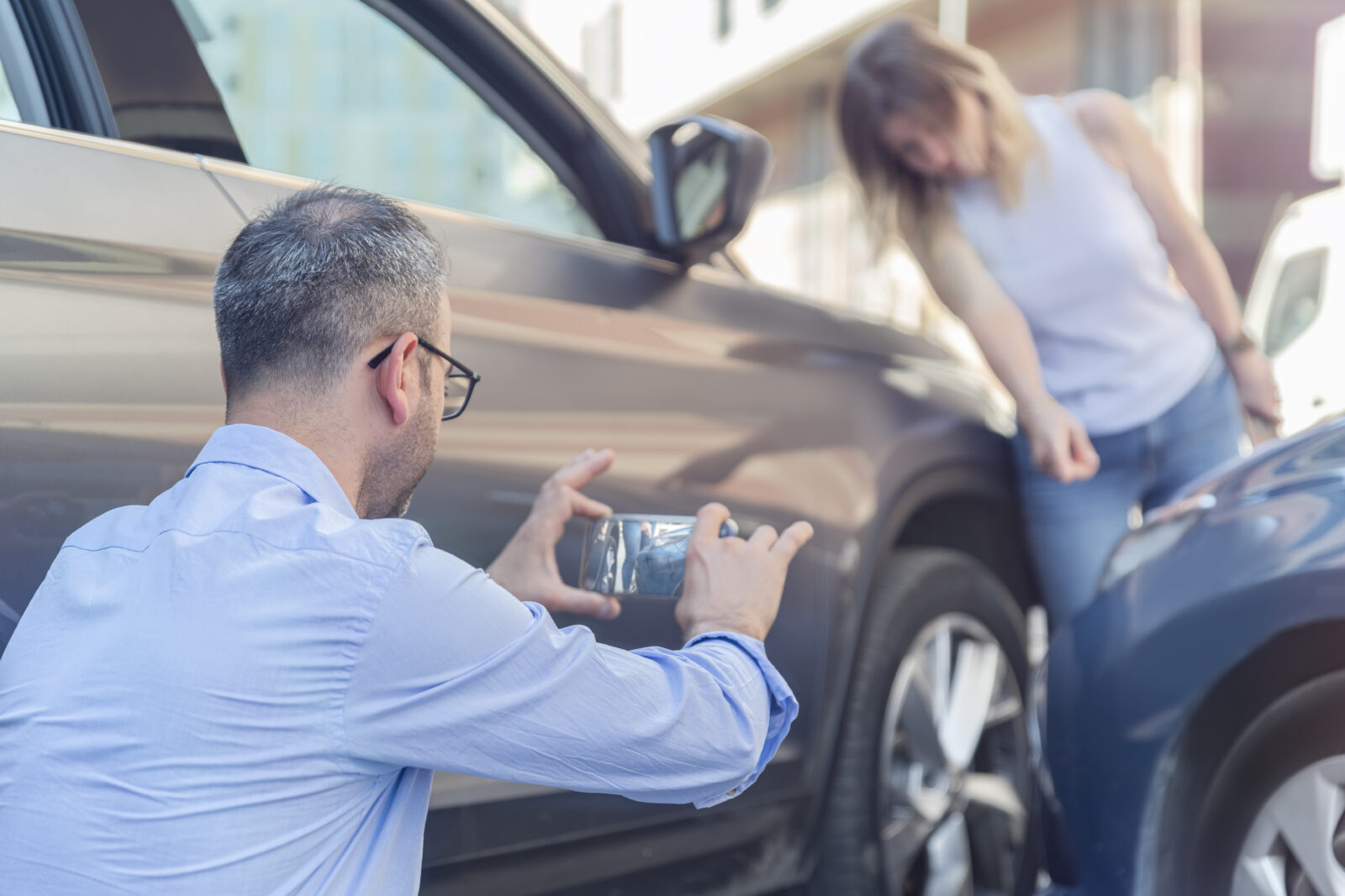 Man taking a photo of vehicle damage with his phone after a car accident, while a woman points at the damage.