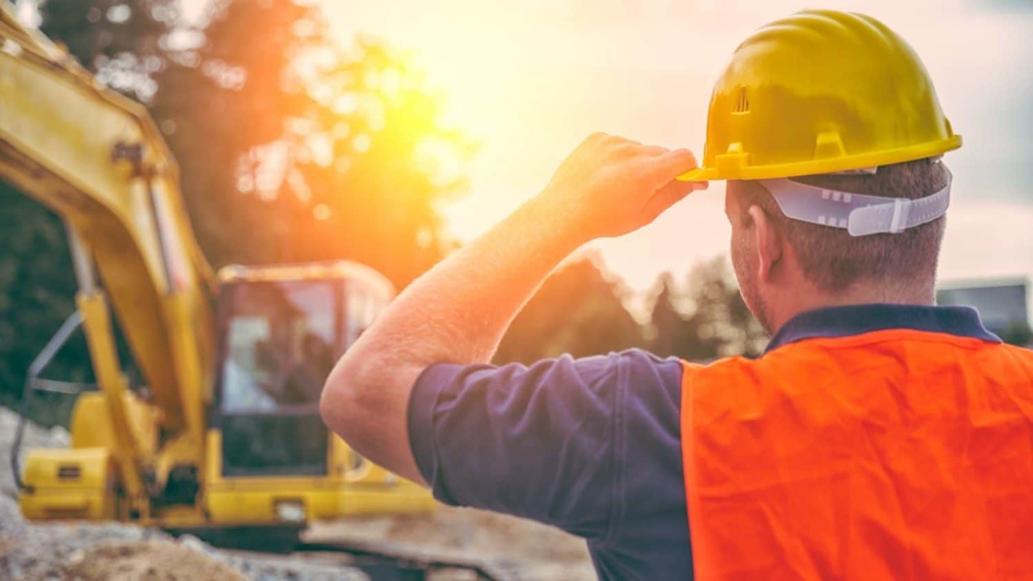 Construction worker in a hard hat and safety vest overlooking an excavator at a job site during sunset.