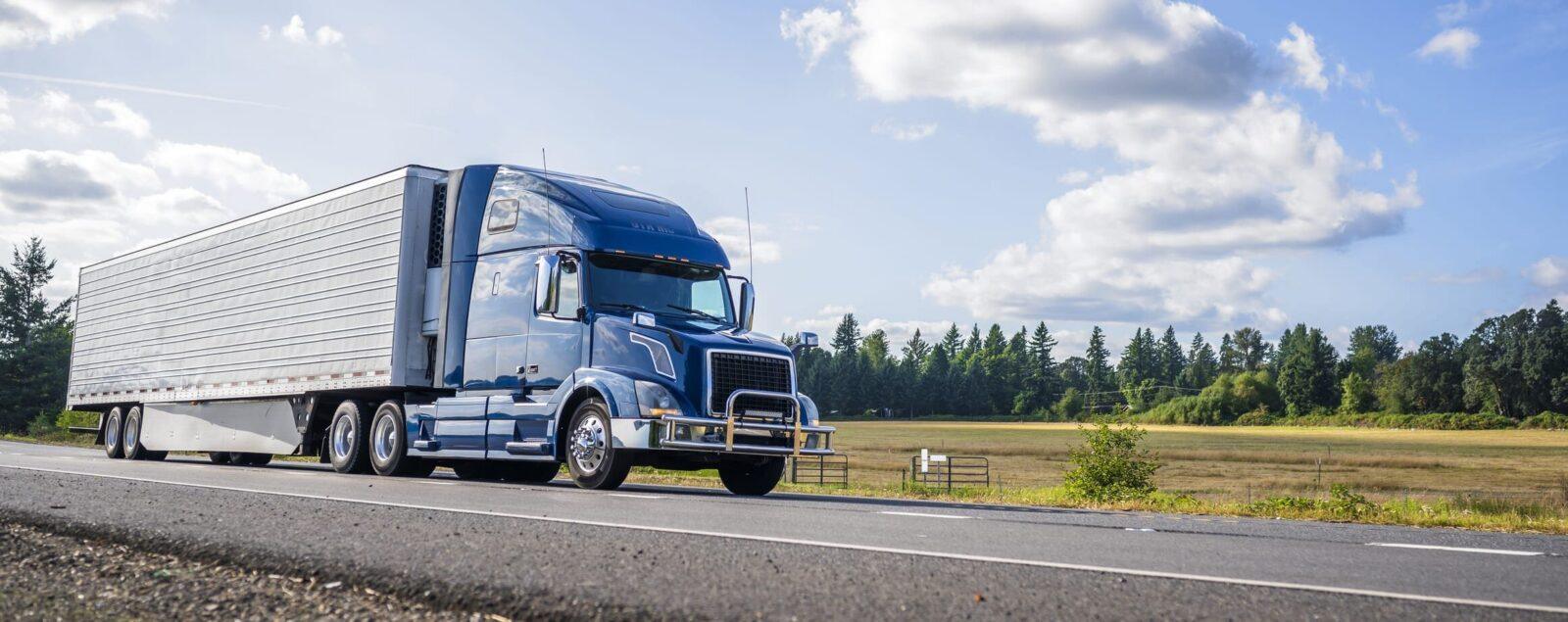 A blue semi-truck driving on a rural highway, with fields and trees lining the road.