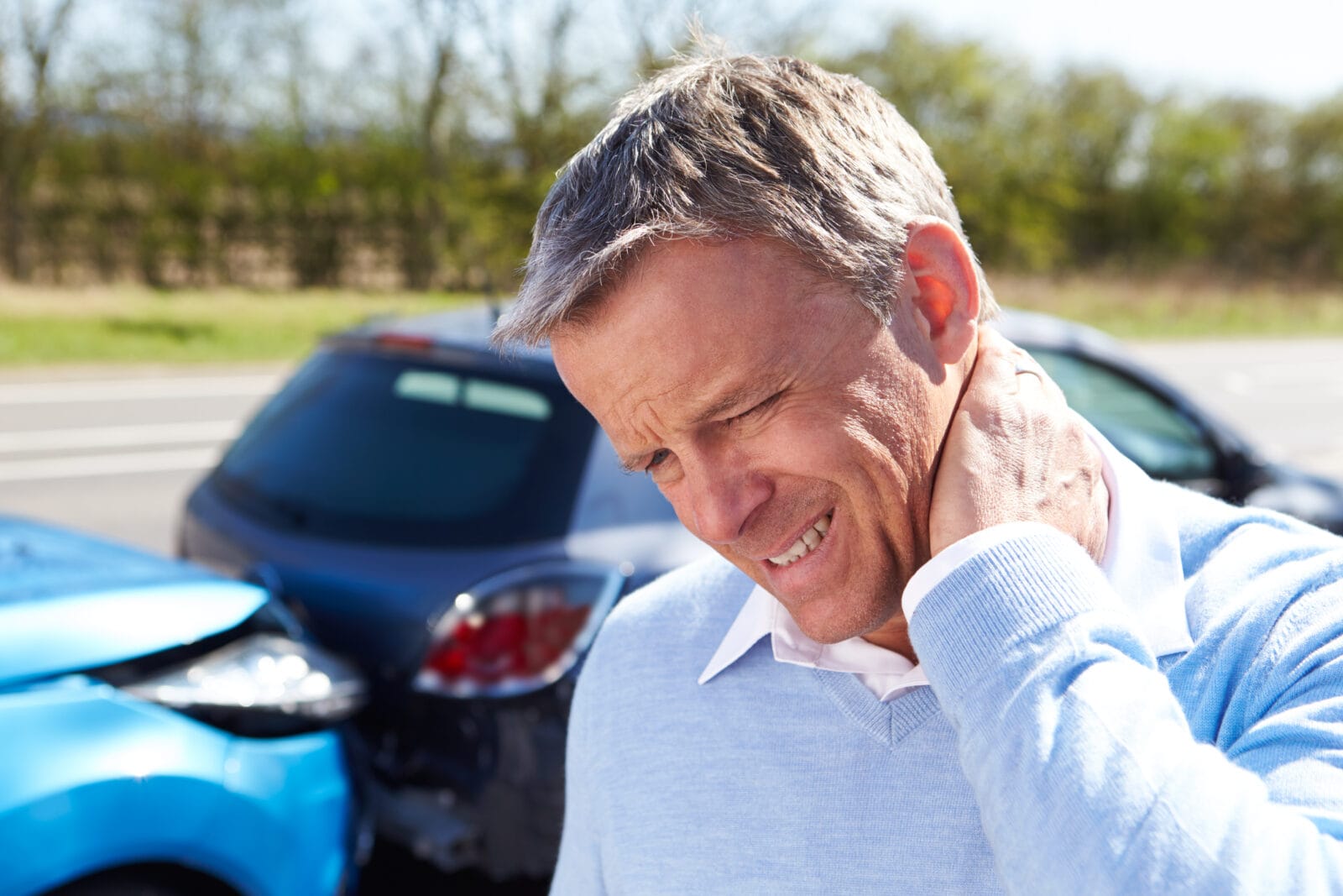 Man holding his neck in pain after a car accident, with two damaged vehicles in the background.