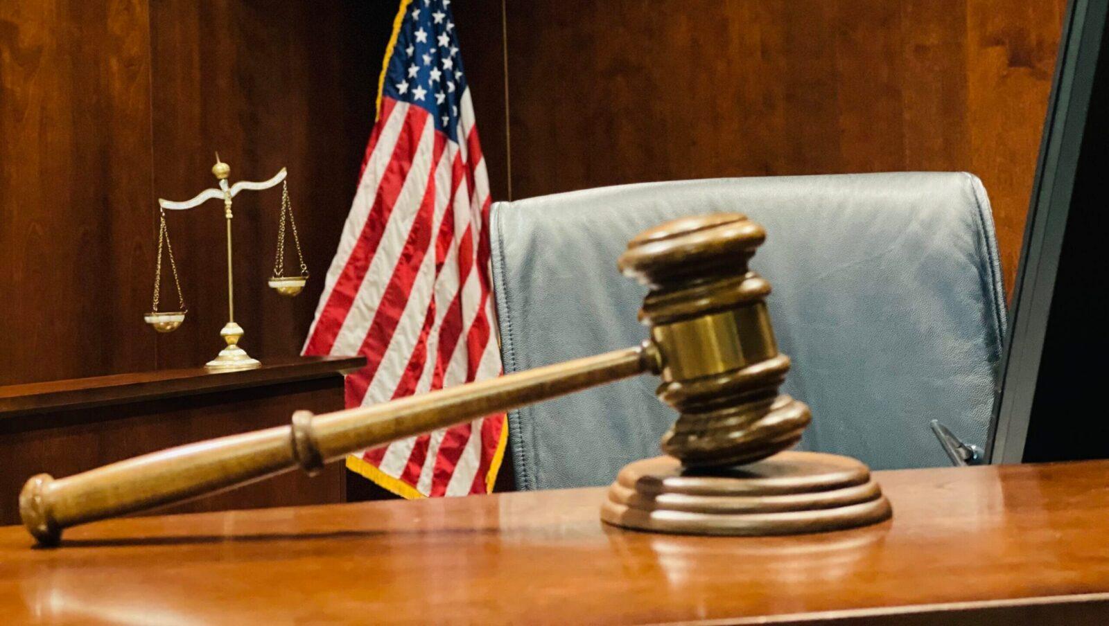 A judge's wooden gavel rests on a sound block at a judicial bench. The scales of justice and an American flag are in the background.