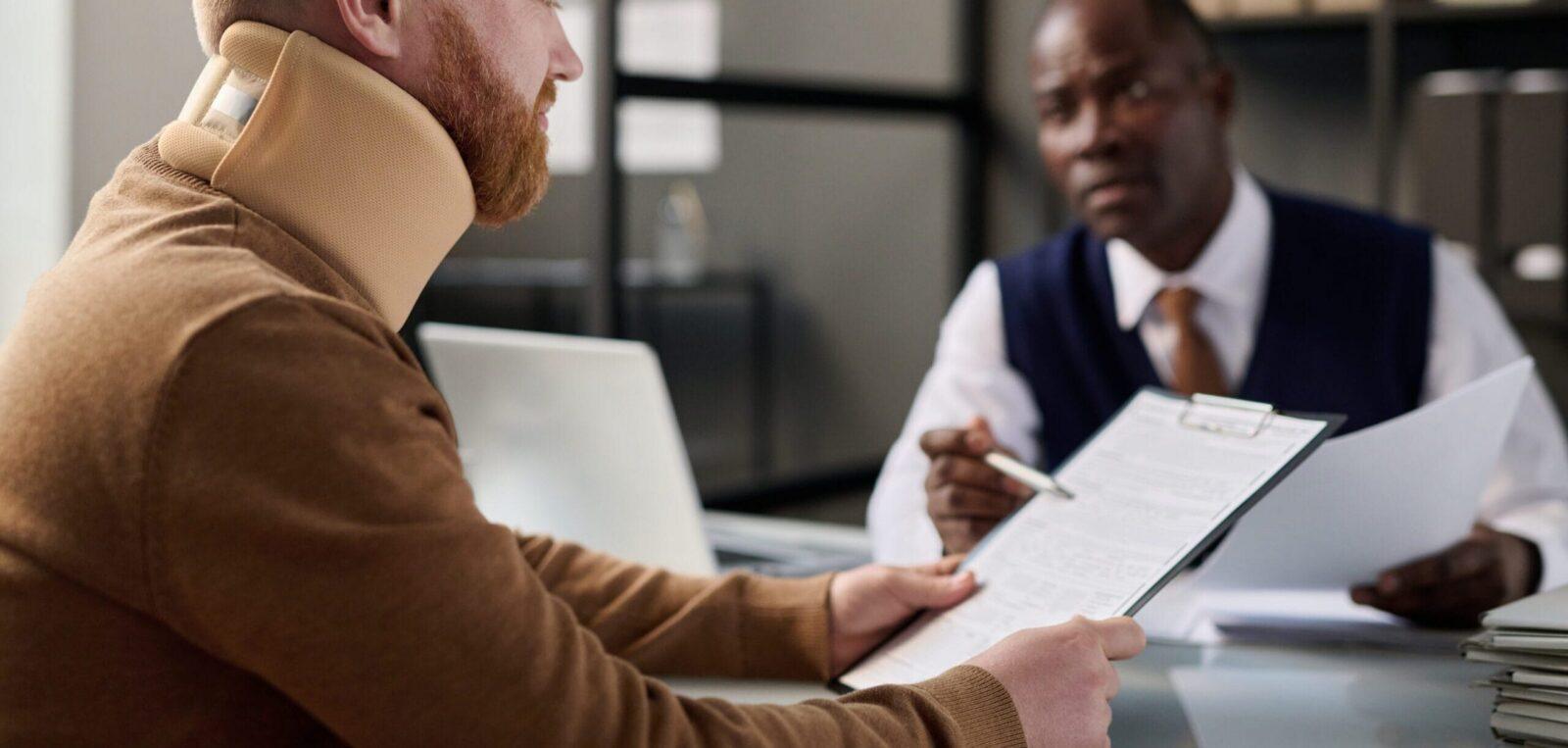 A man wearing a neck brace reviews documents at an office while speaking with a lawyer across the desk.