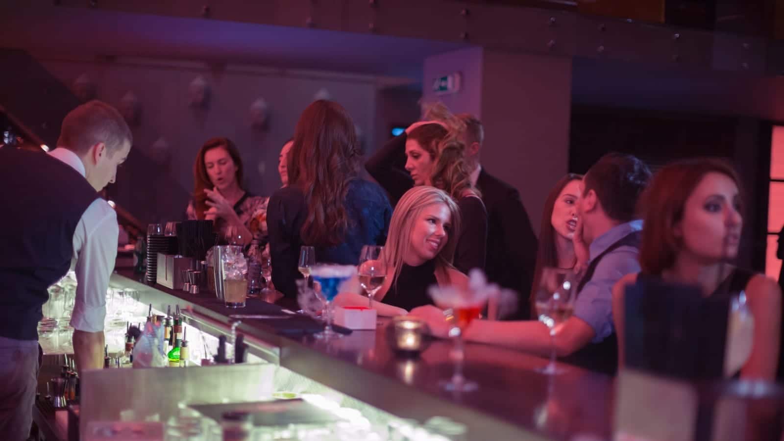 People socializing and drinking cocktails at a busy bar with a bartender serving drinks.