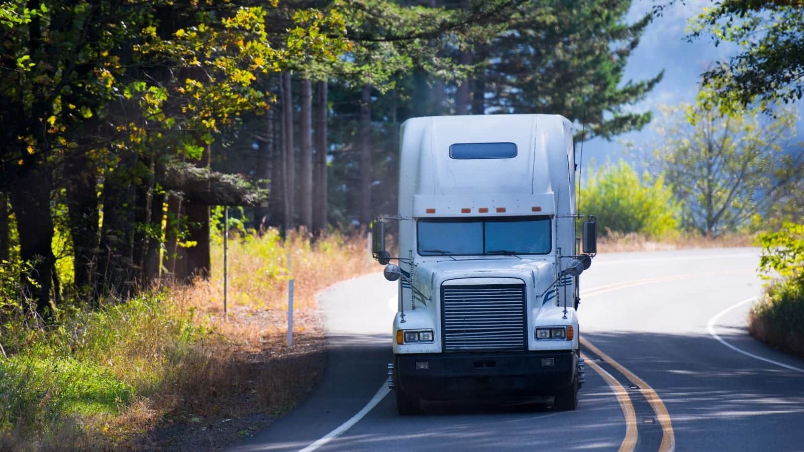 A large white semi-truck driving on a curved two-lane road through a wooded area on a sunny day.